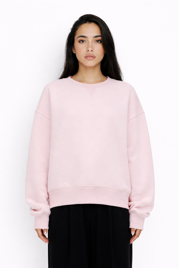 Model trägt rosafarbenes Sweatshirt mit Rundhalsausschnitt und V-Naht am Kragen. Überschnittene Schultern und lange Ärmel mit Rippbündchen. Gerader Saum mit breitem Bund. Locker geschnittene Passform, kombiniert mit schwarzem Rock.