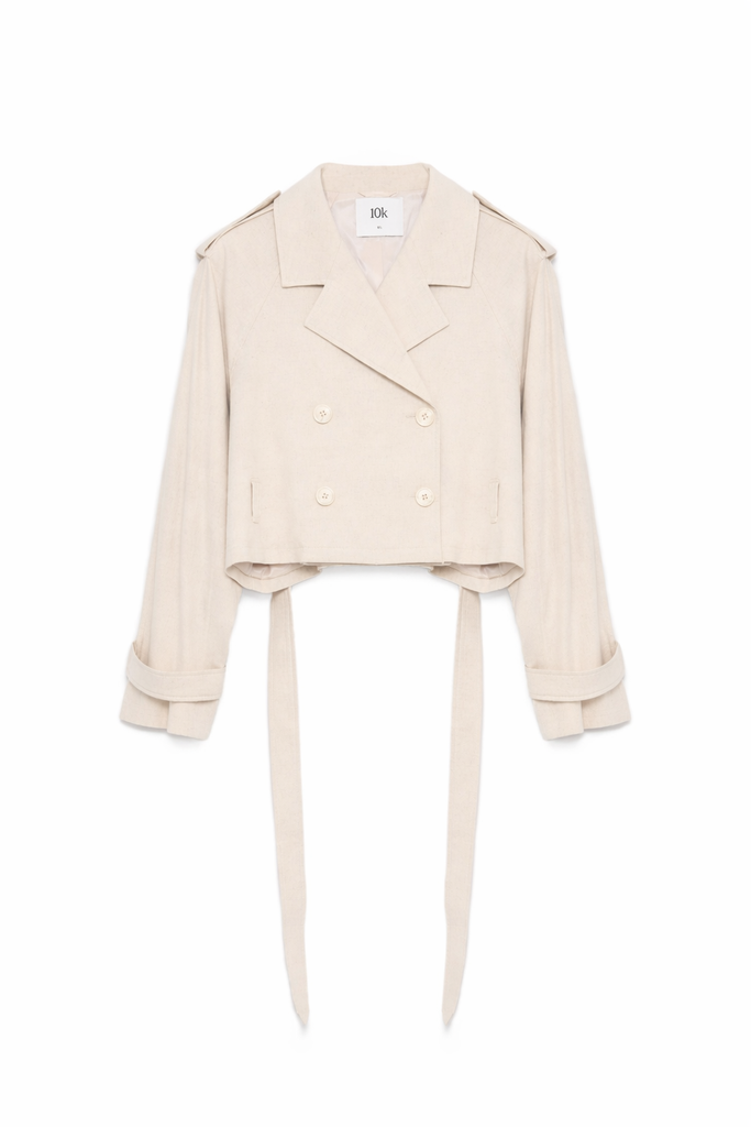 Kurze, cremefarbene Jacke mit Reverskragen, doppelreihiger Knopfleiste, langen Ärmeln mit Riegeln und zwei langen Bindebändern am Saum, freigestellt vor hellem Hintergrund.
